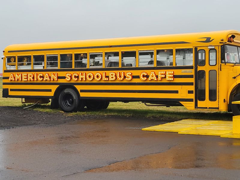 A bright yellow American school bus has been converted into a café, with the words 'AMERICAN SCHOOLBUS CAFE' displayed in large, illuminated marquee-style letters along its side. The bus is parked on a grassy area with a wet, muddy surface in the foreground, and a yellow platform is placed near the entrance. The windows reveal a glimpse of the interior, with tables and seating visible inside. The overcast sky gives the setting a slightly moody atmosphere.