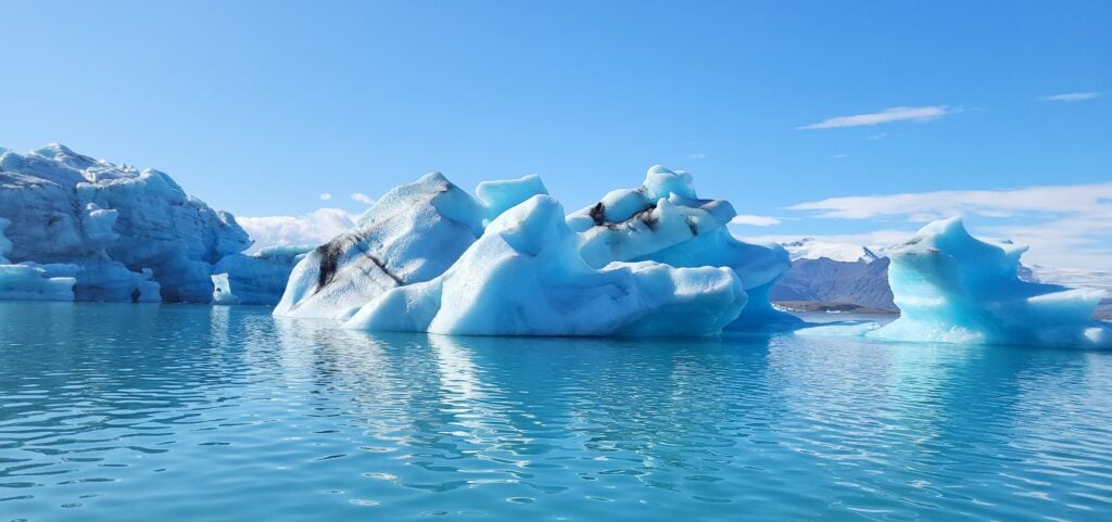 A breathtaking view of Jökulsárlón Glacier Lagoon in Iceland, featuring large, bright blue icebergs floating on calm, crystal-clear water. The ice formations have unique shapes with streaks of black volcanic ash, set against a backdrop of distant mountains and a clear blue sky.