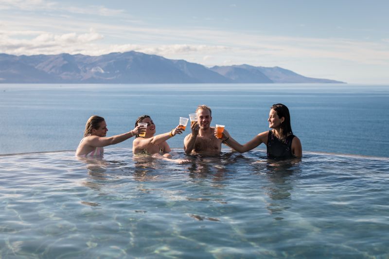 "A group of four people enjoying the geothermal infinity pool at GeoSea in Húsavík, Iceland. They are raising their drinks in a cheerful toast, surrounded by the tranquil blue waters of the pool. The view behind them showcases a breathtaking expanse of the ocean with majestic mountains in the distance. The clear sky and sunlight add to the relaxing and celebratory atmosphere of this scenic spa experience."