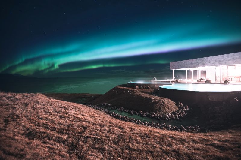 "GeoSea geothermal sea baths in Húsavík, Iceland, illuminated at night under a breathtaking display of the Northern Lights. The infinity pools glow against the dark landscape, reflecting the vibrant green and blue hues of the aurora borealis. The modern architecture of the spa blends seamlessly with the rugged coastal surroundings, creating a serene and magical atmosphere. The ocean stretches into the distance, adding to the sense of tranquility and wonder."