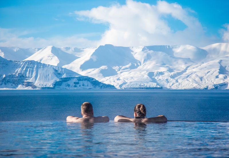 "Two people relaxing in the geothermal infinity pool at GeoSea in Húsavík, Iceland, gazing out at a stunning winter landscape. Snow-covered mountains stretch across the horizon, contrasting with the deep blue waters of the ocean. The bright sky and soft clouds add to the serene atmosphere, highlighting the peacefulness of this unique spa experience, where warm geothermal waters meet the crisp Arctic air."