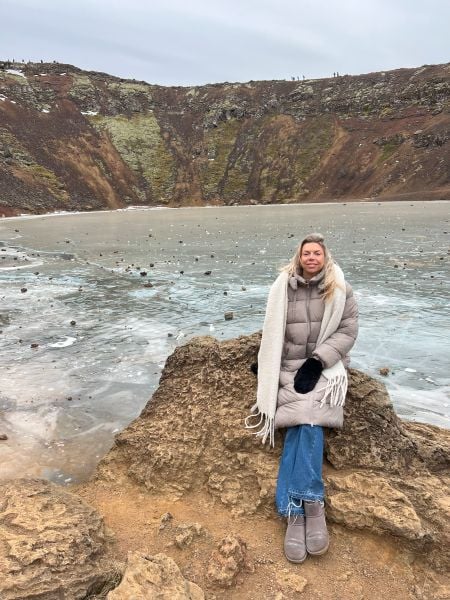 A woman dressed in a beige puffer coat, blue jeans, and gray boots sits on a large rock at the edge of a frozen crater lake. She is wrapped in a long cream-colored scarf, wearing black gloves, and has a relaxed expression as she looks at the camera. The crater’s steep walls in the background have patches of green moss and reddish-brown earth, with small figures of people walking along the rim. The lake’s icy surface is scattered with small rocks, and the overcast sky gives the scene a cool, wintery atmosphere.