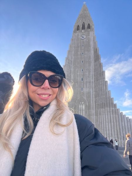 A woman with long blonde hair, wearing a black hat, sunglasses, a black coat, and a cream-colored scarf, smiles while taking a selfie in front of Hallgrímskirkja, the iconic concrete church in Reykjavik, Iceland. The sky is bright blue with a few wispy clouds, and sunlight casts a warm glow on her face. The towering church, with its distinct stepped design, stands prominently in the background, with a few people walking near its entrance.