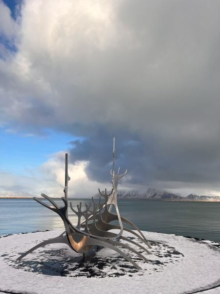 The Sun Voyager (Sólfar) sculpture in Reykjavik, Iceland, stands on a snowy shoreline with the calm sea and distant mountains in the background. The stainless steel structure, resembling a Viking ship, reflects light against a sky filled with a mix of dark clouds and patches of blue. The contrast between the snow-covered ground, the metallic sculpture, and the dramatic sky creates a striking composition.