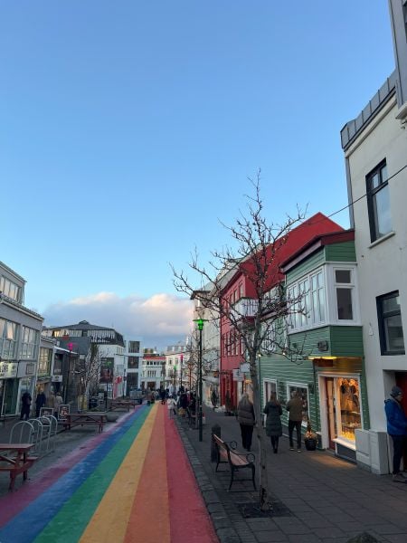 A vibrant street scene in Reykjavik, Iceland, featuring the famous rainbow-painted pavement of Skólavörðustígur leading towards the city center. The street is lined with colorful buildings, including a green and red house with large windows displaying warm lighting. Leafless trees and benches line the sidewalk, and people are strolling along the street, enjoying the shops and cafes. The sky is clear blue with a few soft clouds, and the setting sun casts a gentle glow on the scene.