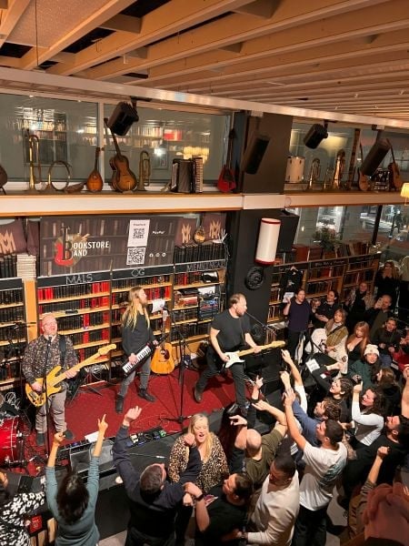 A lively indoor concert at a bookstore-themed venue, where a three-member band is performing on a small stage surrounded by bookshelves. The band members include a guitarist, a bassist with long blonde hair playing a keytar, and a lead singer with a black t-shirt playing an electric guitar. The crowd is packed and enthusiastic, with many raising their hands and singing along. The warm lighting, musical instruments displayed on shelves, and the venue’s cozy yet energetic atmosphere create an intimate live music experience.