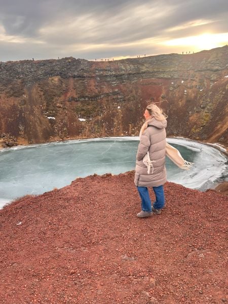 A woman wearing a long beige puffer coat, blue jeans, and brown boots stands on the edge of a crater, gazing at a frozen lake below. Her blonde hair is blowing in the wind, and a cream-colored scarf flutters behind her. The crater walls are reddish-brown with patches of snow, contrasting with the icy blue surface of the lake. The sky is cloudy with a warm glow near the horizon, and small silhouettes of people can be seen walking along the crater’s rim in the distance.