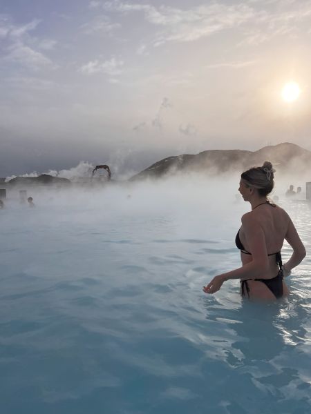 A woman in a black bikini stands partially submerged in the milky blue waters of the Blue Lagoon in Iceland. Steam rises from the geothermal pool, creating a misty atmosphere. The sun is low in the sky, casting a soft golden glow over the scene, with distant hills and other bathers visible through the haze. The tranquil water and mist blend with the overcast sky, adding to the spa’s serene and otherworldly ambiance.