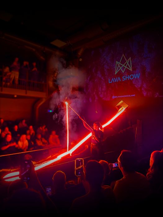 This is a scene from Lava Show. A woman wearing glasses, a headset, and a red shirt is demonstrating molten lava in a dramatic indoor setting. She is holding a long metal rod with a glowing, fiery-hot substance at the end, creating a trail of light as she lifts it. The scene is illuminated by the intense orange and red glow of the molten lava, reflecting off the surrounding rocky structures. A group of spectators watches from a balcony in the dimly lit background, adding to the immersive, volcanic atmosphere.