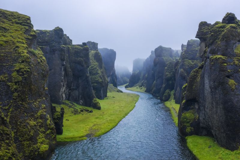 A winding river flows through the dramatic Fjaðrárgljúfur canyon in Iceland, surrounded by towering, moss-covered cliffs. The steep rock formations, shaped by centuries of erosion, rise on either side of the water, creating a breathtaking, otherworldly landscape. A soft mist lingers in the background, adding to the mysterious and serene atmosphere. The vibrant green moss contrasts with the dark, rugged rock, enhancing the canyon’s striking natural beauty.