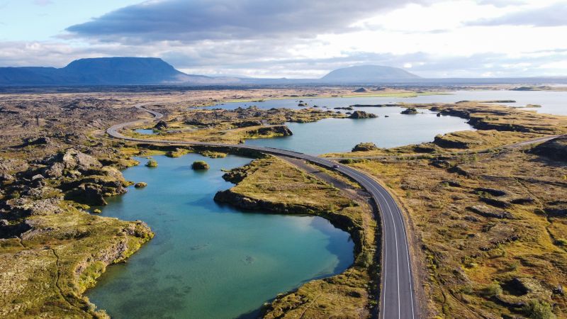 A winding road curves through the volcanic landscape of Lake Mývatn in Iceland, surrounded by vivid blue waters and rugged lava formations. Distant mountains rise under a partly cloudy sky, adding to the dramatic scenery.