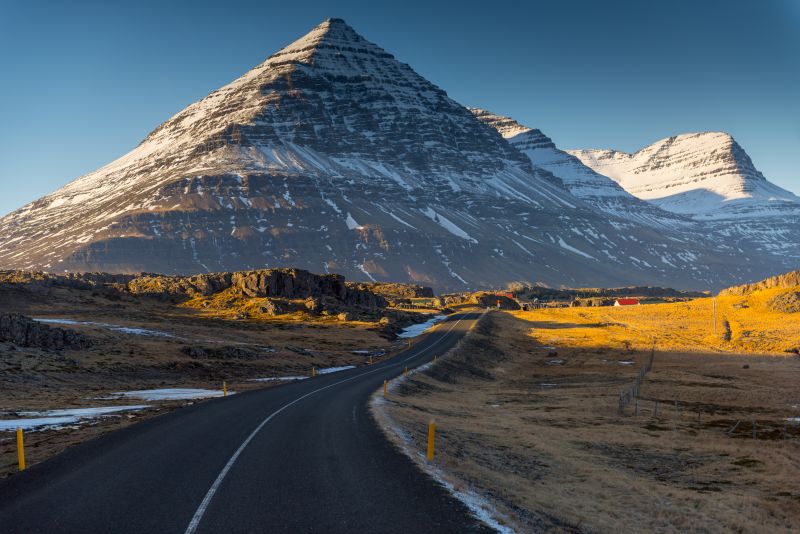 A winding asphalt road with yellow markers curves through a rugged Icelandic landscape, leading toward a towering, snow-capped mountain with steep, layered slopes. The late afternoon sunlight casts a golden glow on the surrounding grassy fields and rocky terrain, contrasting with the cool tones of the mountain and the deep blue sky. In the distance, a few red buildings stand out against the dramatic backdrop, adding a sense of scale and isolation to the remote setting.