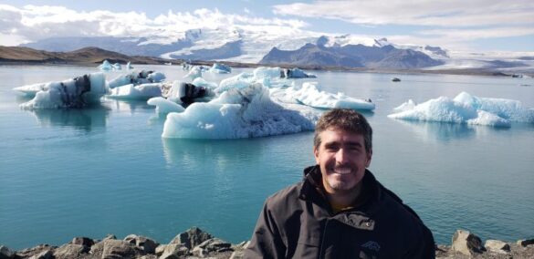 A man with short dark hair and a beard, wearing a black jacket, smiles at the camera while standing in front of Jökulsárlón Glacier Lagoon in Iceland. Behind him, the lagoon’s calm, icy blue waters are dotted with floating icebergs of various shapes and sizes. Some of the icebergs have streaks of black from volcanic ash. In the background, rugged mountains and a massive glacier stretch across the horizon under a partly cloudy sky. A small boat is visible in the distance on the water.